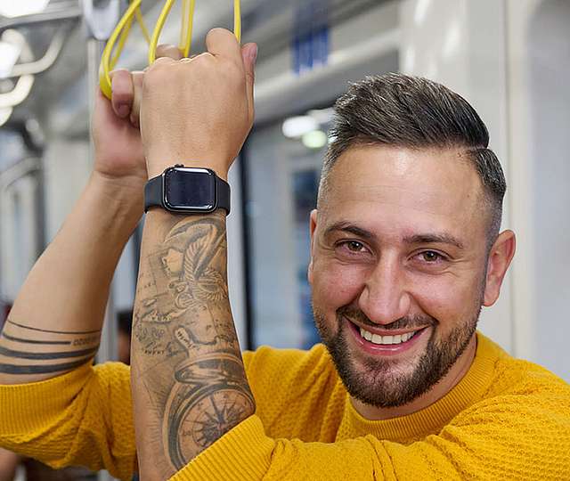 Ein lächelnder Mann steht in der Straßenbahn und hebt sich mit beiden Händen an einem Haltegriff fest